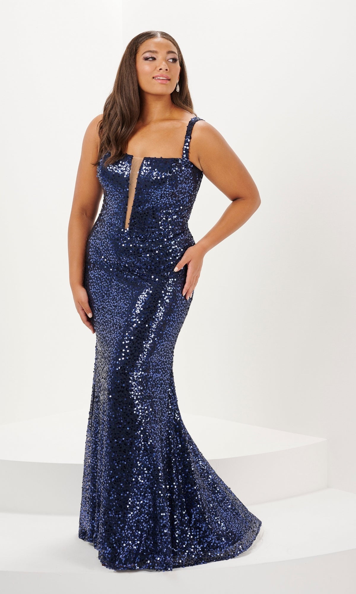 A woman stands on a white platform wearing the Tiffany Long Plus-Size Sequin Prom Dress 16123— a sleeveless navy blue mermaid gown with an illusion plunging neckline, set against a plain white background.